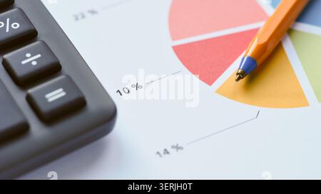 An orange pen lying on a colorful pie chart document next to a black office calculator, symbolizing financial planning and business analysis. Close up Stock Photo