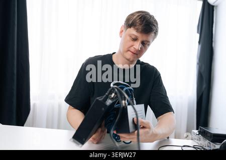 Focused man in black t-shirt sitting at white table, examining and assembling black electronic equipment with tangled blue wires. Bright background Stock Photo