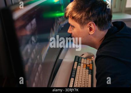 Profile view of young man concentrating intently on curved computer screen. He leaning very close, face illuminated by monitor light in dimly lit room Stock Photo