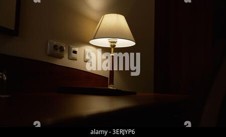 Soft warm light emanates from table lamp, casting gentle glow on textured wall electrical outlets. Cozy interior setting creates peaceful intimate moo Stock Photo
