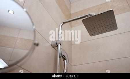 Contemporary chrome shower fixture featuring square rain shower head attached to curved arm mounted on beige ceramic wall tiles. Bright lighting highl Stock Photo