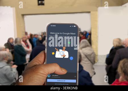 Hand holding a smartphone displaying Google Meet app with a blurred crowd of people & spectators in the background Stock Photo