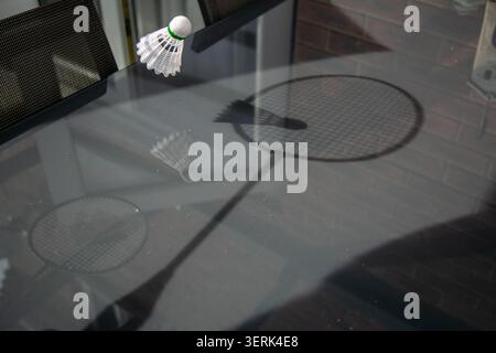 White badminton shuttlecock with a green band placed on a glass table, casting the shadow of a badminton racket beside it. Stock Photo