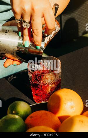 Woman pours drink into glass with mixed fruits nearby on a sunny day in a casual outdoor setting. girl holds cocktail shaker and pours  red drink into Stock Photo