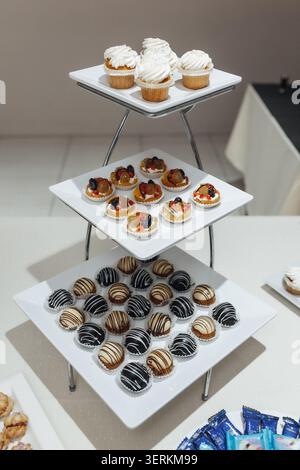 Three-tiered stand holds assorted pastries. White plates display cupcakes, tarts, and chocolate-covered treats. Sleek silver legs support elegant dess Stock Photo