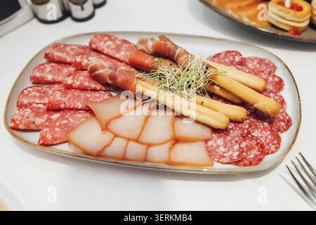 Platter holds assorted meats and asparagus. Salami slices, prosciutto-wrapped sticks, and fresh sprouts. Delicate presentation on a white tablecloth. Stock Photo