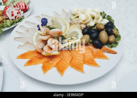 Fresh seafood platter gleams on a crisp white plate. Shrimp, salmon, olives, and lemon slices arranged artfully. Bright colors contrast with soft, ele Stock Photo