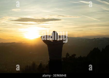 Silhouetted figure stands atop hill at golden hour. Hands clasped behind head, eyes closed in peaceful stillness. Sun dips behind distant mountains, c Stock Photo