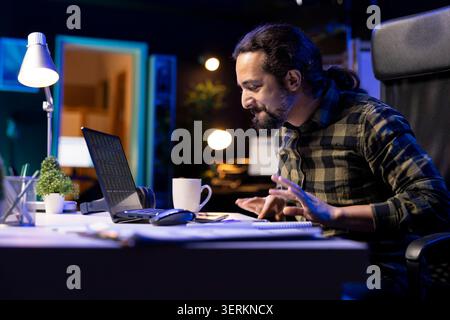 In his comfortable apartment, bearded man diligently works from home on his laptop, focused on financial research. Male freelancer carefully analyzes investment charts and studies market trends. Stock Photo