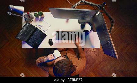 Top-down view of self employed man doing IT support job from apartment personal office, coding on PC, drinking coffee. Software developer writing lines of code on computer at home, aerial shot. Stock Photo