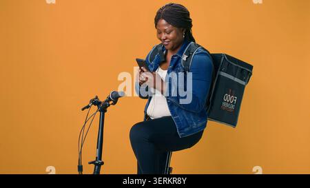 Healthy African American female with bag on her back using mobile communication for on-the-go urban delivery service, active black woman happily receiving online orders on cell-phone from customers Stock Photo