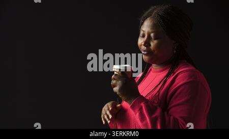 African American woman immersed in pleasure of sipping coffee lost in contemplation gazing upwards, female fashion blogger savors her warm beverage with sense of satisfaction, side-view portrait Stock Photo