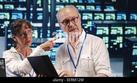 Software developers in high tech establishment, completing tasks on tablet while running scripts on laptop terminal, proactive programmers amidst server rows. Stock Photo