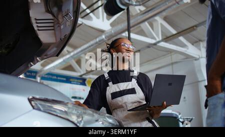 Experienced technician listens to customer in repair shop, using laptop to write down car modifications needed, professional in garage with client, listening to her requests, zoom out shot. Stock Photo
