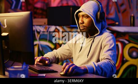 Hacker stealing credit card numbers and manipulating banking systems for his own personal gain while listening to music through headphones, evil developer programming in graffiti painted basement. Stock Photo