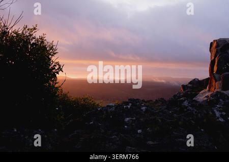 Sunset paints distant hills in warm orange glow. Dark rocks and shrubs frame the view at a peak near Cradle Mountain in Tasmania, Australia. Stock Photo