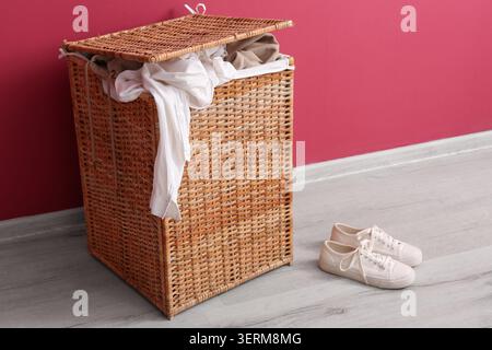 Wicker laundry hamper with dirty clothes and white sneakers on wooden floor against pink wall, common household chore and the need for organization in Stock Photo