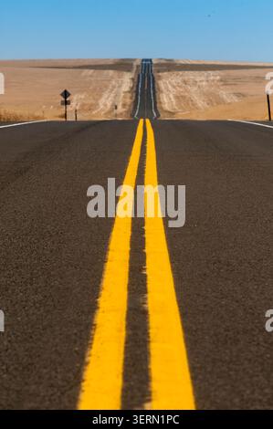 Straight two-lane highway with bold yellow centerlines stretches endlessly through golden wheat fields under a clear blue summer sky. Stock Photo