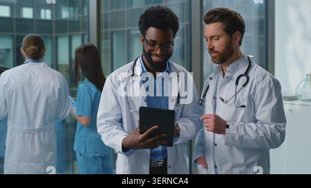 Two multiracial doctors men talking with digital tablet teamwork in clinic. African American practitioner with Caucasian surgeon specialist smiling Stock Photo