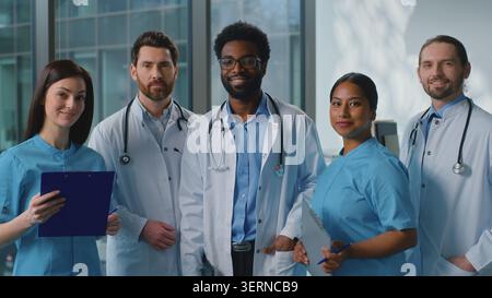 Happy smiling multiethnic doctors look in digital tablet talking discussing online health test results in clinic. Joyful team five surgeons looking at Stock Photo