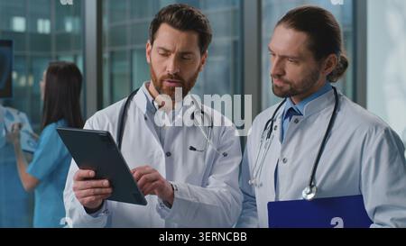 Two Caucasian doctors men talking analyzing health checkup results with digital tablet online medicine service in clinic. Coworkers physicians Stock Photo