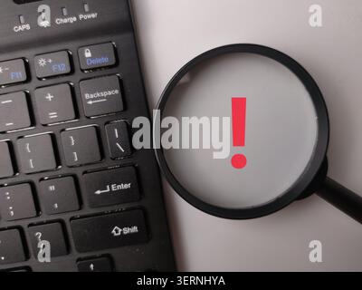 A magnifying glass placed over a white background highlights an overlaid red exclamation mark icon next to a keyboard. Stock Photo