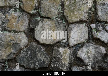Rough stone wall with mossy, weathered surfaces. Grey stones vary in size and shape. Some stones show signs of age and wear. Moss grows in patches amo Stock Photo