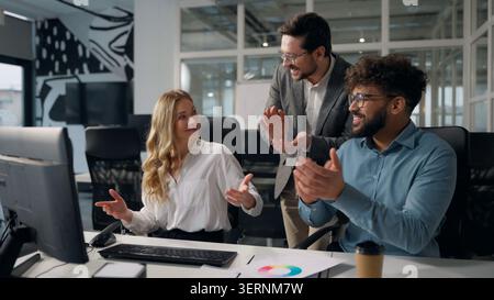Three happy multiethnic business colleagues working laptop together applauding good result winning online applause team Caucasian businesswoman typing Stock Photo