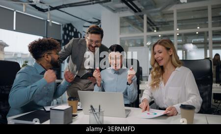 Four multiethnic business people working together with laptop talking winning job success giving high five celebrating multiracial businessmen and Stock Photo