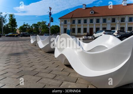 Modern white wave-shaped benches on a paved sidewalk in a bright urban setting with a classic building and blue sky background. Stock Photo