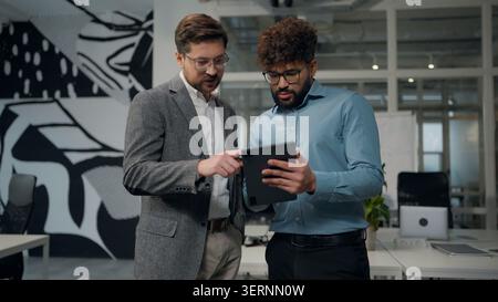 Two successful multiethnic business men talking looking in digital tablet gadget device using together in office diversity businessmen talk analyzing Stock Photo