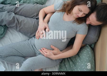 Expecting mother and partner lying in bed with hand on belly and arm around shoulder. Pregnant woman resting head on husband chest warmly. Mom-to-be and man enjoying quiet intimate morning at home Stock Photo
