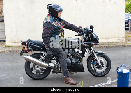 A man wearing a helmet & a denim vest with patches sits on a black Yamaha Fazer motorcycle on a street Stock Photo