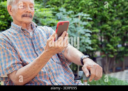 Happy senior man with walking cane using his smart phone in the park. Wellness of old person, walking stick and support concept. Stock Photo
