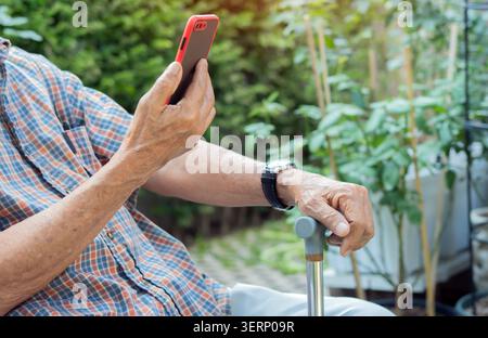 Side view of unrecognizable elderly man sitting at park with walking stick and using his smart phone. Retirement technology lifestlye concept. Stock Photo