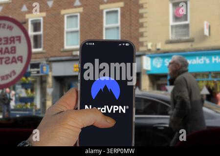 Hand holding a smartphone displaying the NordVPN logo on the screen with a blurred UK high street in the background Stock Photo