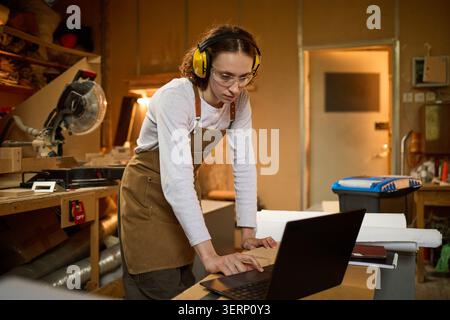 Adult Woman Carpenter Using Laptop in Workshop for Project Planning Stock Photo