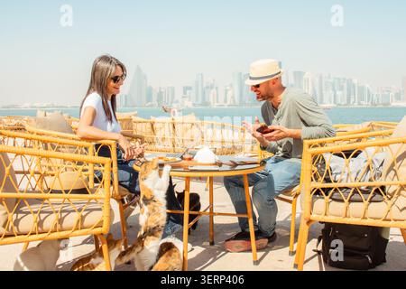 Close up Man and woman sitting in outdoor cafe Doha Corniche. Woman playing feeding cats, man using smartphone having fun. Wicker chairs, table, food, Stock Photo