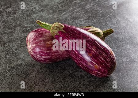 Two fresh purple striped eggplants resting on dark textured stone background. Vibrant purple skin with white streaks green stems. Healthy organic vege Stock Photo