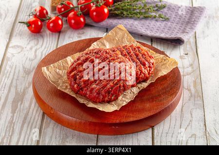 Uncooked beef burger patties on parchment paper, rustic wooden cutting board. Fresh red cherry tomatoes green thyme sprigs in background Stock Photo