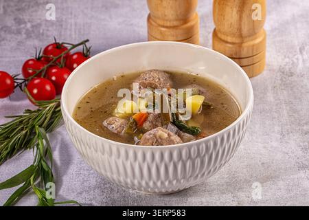Close up of homemade meatball soup with potatoes, carrots, and green beans in rich broth. White ceramic bowl on gray table with fresh tomatoes and her Stock Photo