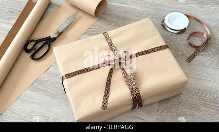 Gift-wrapped package with brown paper and leopard print ribbon on wooden surface, scissors and wrapping paper visible in the background Stock Photo