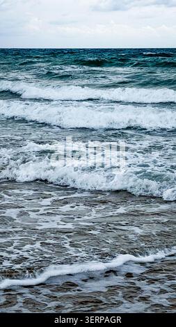 Ocean waves crashing on sandy shore under cloudy sky, with varying shades of blue and white creating a dynamic coastal scene Stock Photo