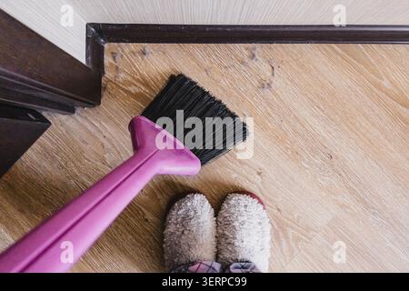 Pink plastic brush with black bristles sweeping dust debris from light wooden laminate flooring, white fluffy slippers visible at bottom, top down vie Stock Photo