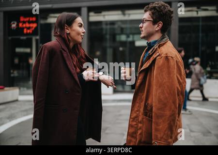 Two colleagues having a winter business meeting outdoors with coffee and coats in an urban plaza Stock Photo