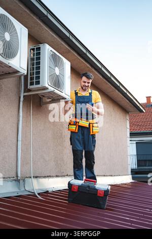 HVAC technician checking air conditioning outdoor unit on the roof using a digital multimeter and smartphone Stock Photo