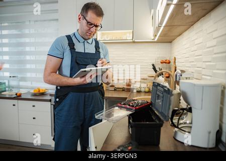 Handyman in overalls writing on a digital tablet while checking a kitchen Stock Photo