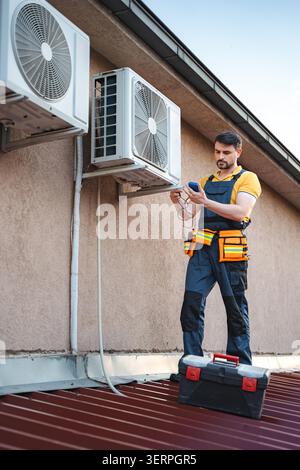 Air conditioner technician installing an air conditioning unit on a roof and checking it with a multimeter Stock Photo