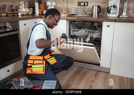 Smiling technician repairing dishwasher using electric screwdriver in modern kitchen Stock Photo
