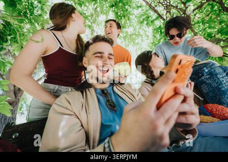 High school students laughing and studying together outdoors while looking at a smartphone under trees Stock Photo